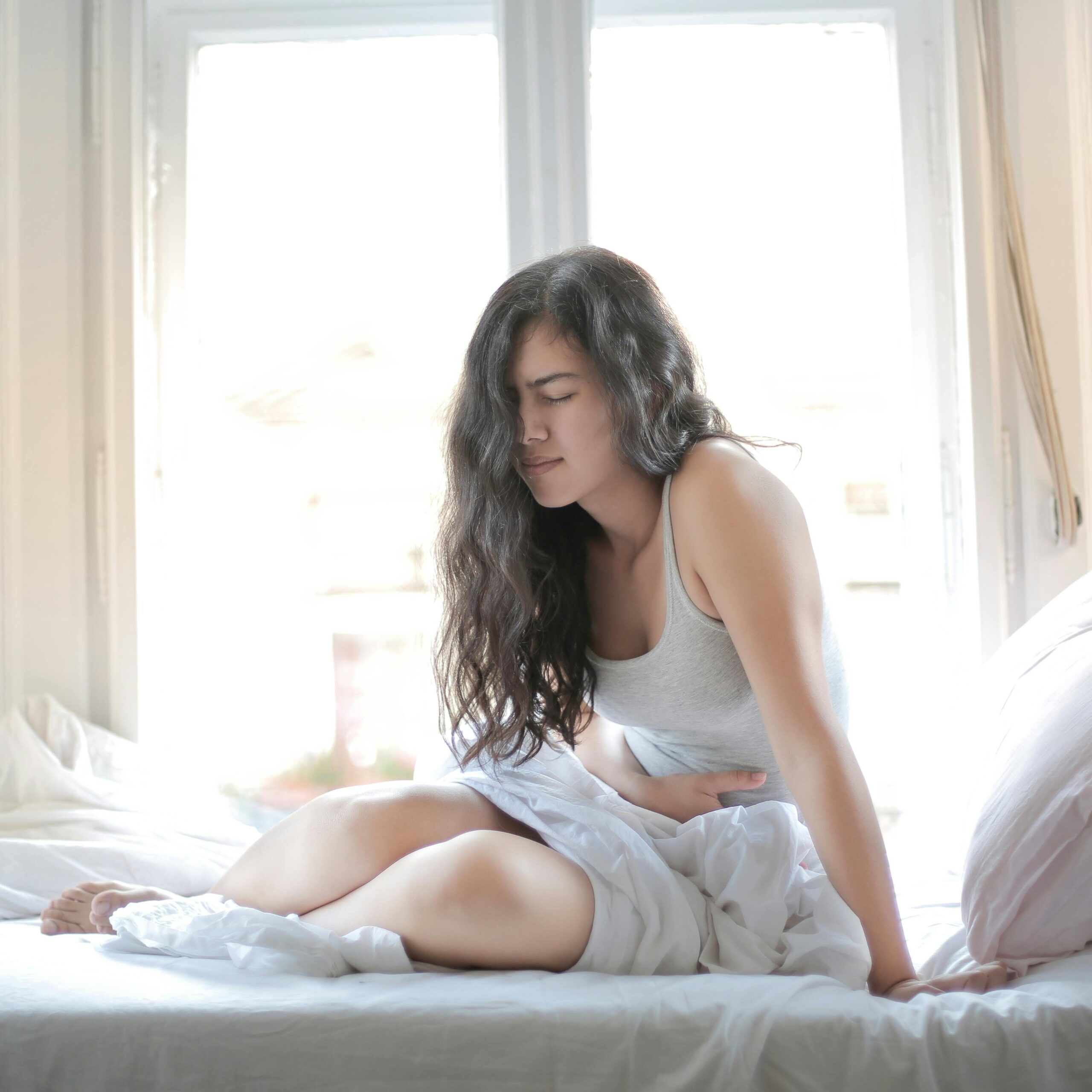 Woman sitting on bed holding her stomach, showing signs of discomfort or pain indoors.